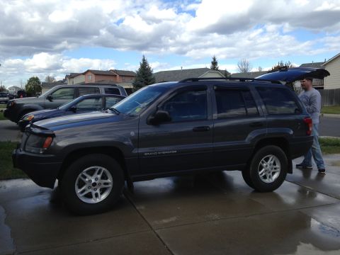2004 Jeep Grand Cherokee - A dark gray 2004 Jeep Grand Cherokee is parked in a driveway with its rear hatch open, alongside other vehicles.