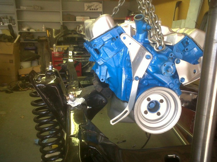 Blue engine block with silver pulley and mounting brackets, suspended by chains in a workshop