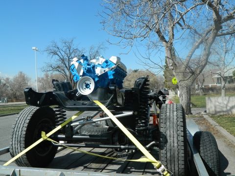 1979 Ford Bronco - A vehicle chassis with a blue and white engine is secured with yellow straps on a trailer, ready for transport.