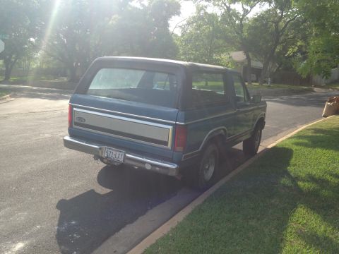 1984 Ford Bronco - Rear view of a blue SUV parked on a street with trees and grass. The vehicle has a chrome bumper and a silver trim panel on the tailgate.