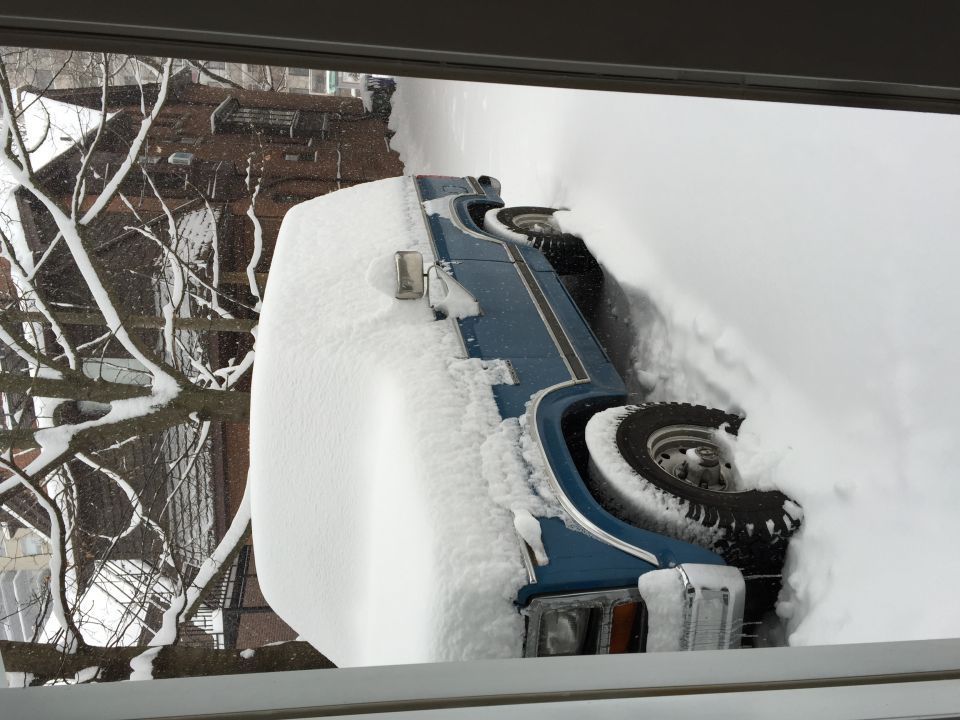 Blue SUV, partially buried in snow, side view, with snow-covered trees and building in background