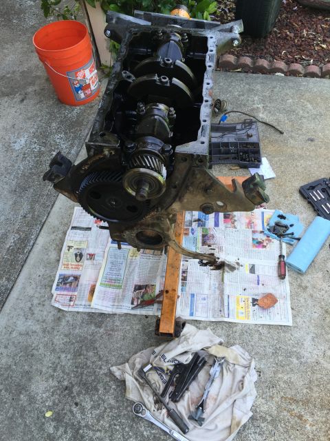 1946 Jeep Willys - An engine block, crankshaft, and gears are laid out on newspapers with tools and an orange bucket, suggesting a repair or rebuild in-progre…