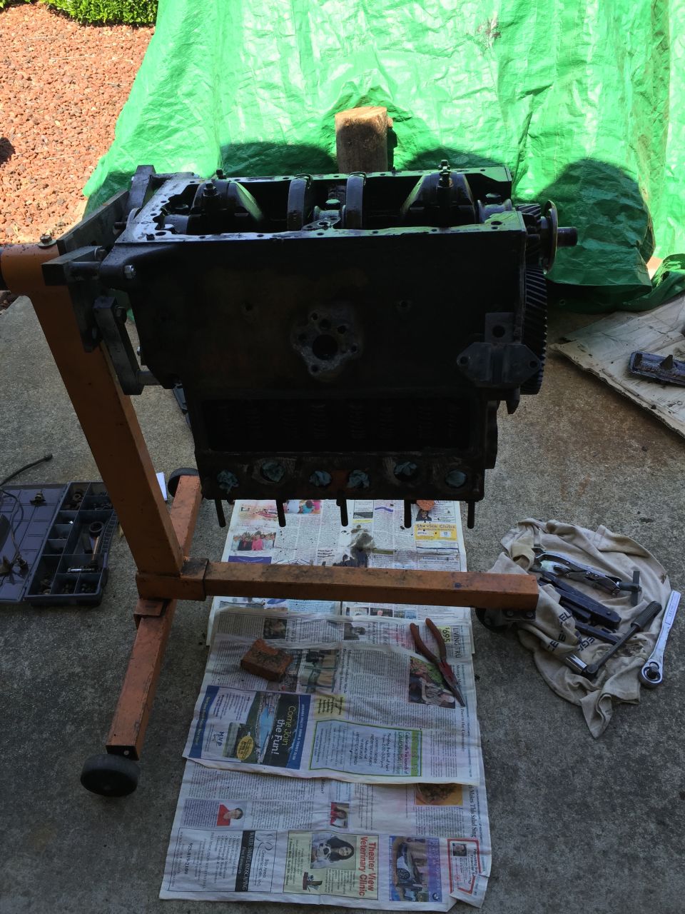 engine block on an orange engine stand, covered in oil, with tools and newspapers underneath