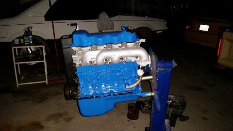1985 Ford Escort - A blue and silver engine is mounted on a stand, with a white vehicle partially visible behind it.