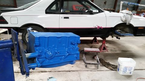 1985 Ford Escort - A white 1985 Ford EXP coupe is elevated on jack stands, with a freshly painted blue engine block positioned in front of it.