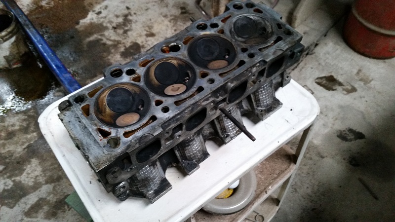 Engine cylinder head with four combustion chambers, resting on a white surface. A blue tool is visible in the background
