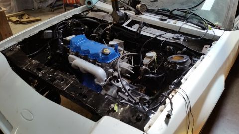 1985 Ford Escort - The engine bay of a car is shown, with a blue valve cover and silver intake pipe.