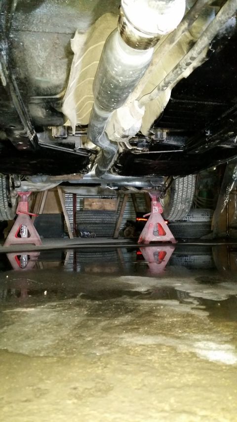 1985 Ford Escort - The underside of a vehicle is shown, supported by jack stands, with a driveshaft and exhaust components visible.