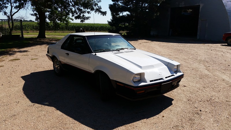 White coupe with a hood scoop parked on a dirt lot
