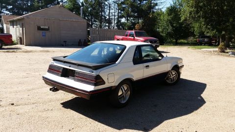 1985 Ford Escort - This two-tone coupe features a prominent rear spoiler and distinctive body lines, suggesting a model year around 1985.