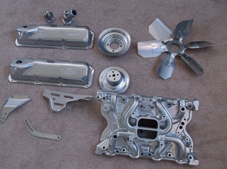 Engine parts including valve covers with Ford logos, a fan, and an intake manifold, laid out on a carpeted surface