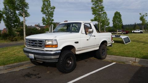 1993 Ford Bronco - A white 1993 Ford Bronco is parked in a lot with its removable top set aside on the grass.