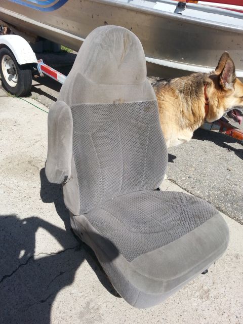 1981 Ford Bronco - A single gray fabric car seat with an armrest sits on pavement next to a boat trailer and a dog.