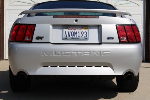 2002 Ford Mustang - Silver Ford Mustang GT with a spoiler and dual exhaust pipes is parked in front of a garage door.