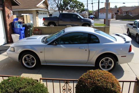 2002 Ford Mustang - A silver coupe with a hood scoop and spoiler is parked in a residential driveway, with a dark pickup truck behind it.
