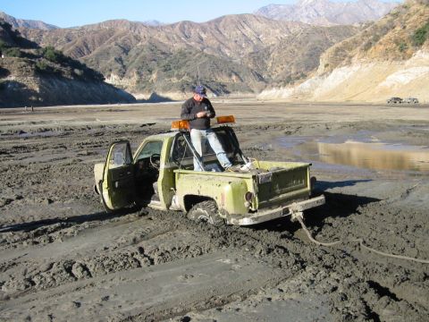 1985 Ford Bronco - A man stands in the bed of a light green 1985 pickup truck that is stuck in thick mud, with mountains in the background.