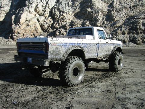 1985 Ford Bronco - This lifted pickup truck is covered in mud, seen from a rear 3/4 angle on a dirt terrain.