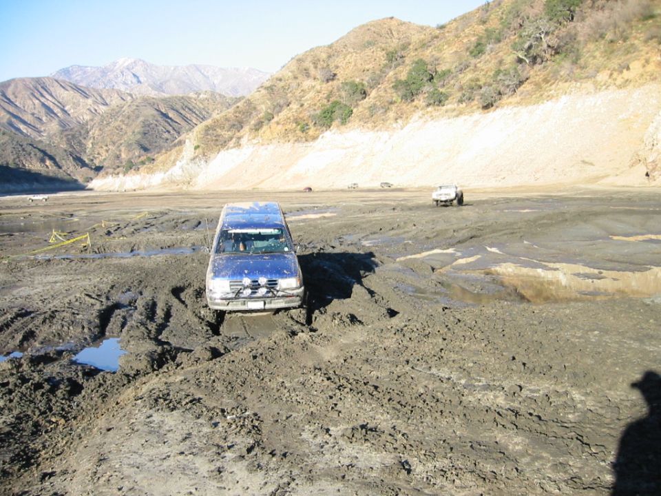 Blue SUV stuck front-first in deep gray mud with hills behind