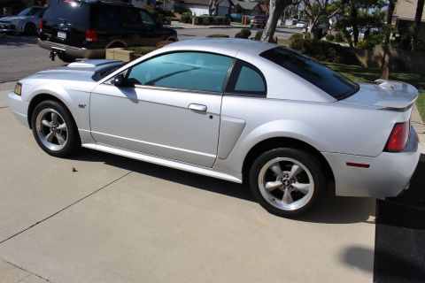 2002 Ford Mustang - A silver coupe with tinted windows and a spoiler is parked on a driveway, with a black SUV and another car in the background.