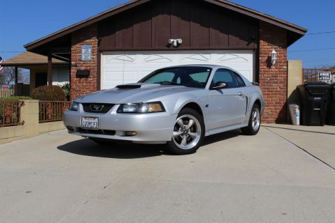 2002 Ford Mustang - A silver Ford Mustang is parked in front of a brick garage on a sunny day. The car has a hood scoop and aftermarket wheels.