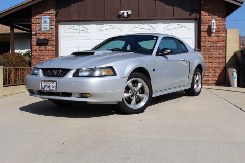 2002 Ford Mustang - A silver Ford Mustang is parked in front of a brick house and garage.