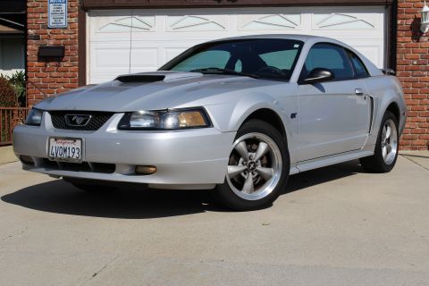 2002 Ford Mustang - This silver 2002 Ford Mustang coupe is captured in a three-quarter front view, parked in front of a brick building with a garage door.