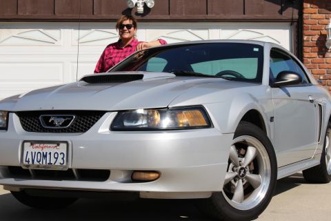 2002 Ford Mustang - A silver 2002 Ford Mustang is parked in front of a garage, with a woman standing next to it.