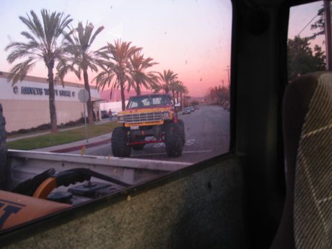 1985 Ford Bronco - A lifted pickup truck with oversized tires drives down a street lined with palm trees at sunset.