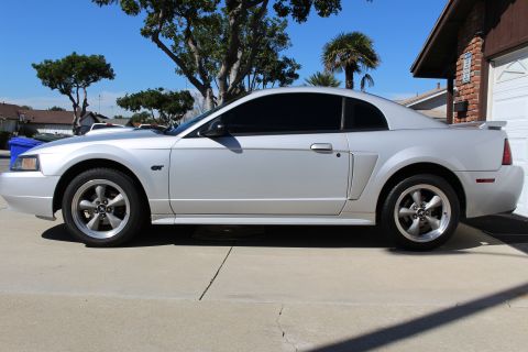 2002 Ford Mustang - A silver 2002 Ford Mustang GT is shown from the side, parked in front of a brick house.