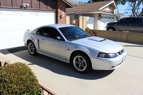 2002 Ford Mustang - A silver 2002 Ford Mustang is parked in a driveway next to a garage, with an SUV visible in the background.