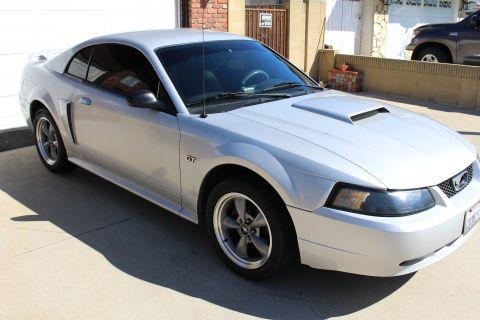 2002 Ford Mustang - Silver 2002 Ford Mustang GT coupe with a hood scoop and GT badging is parked outside a garage.