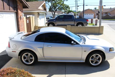 2002 Ford Mustang - A silver coupe is parked in a driveway next to a house, with a gray pickup truck behind it.