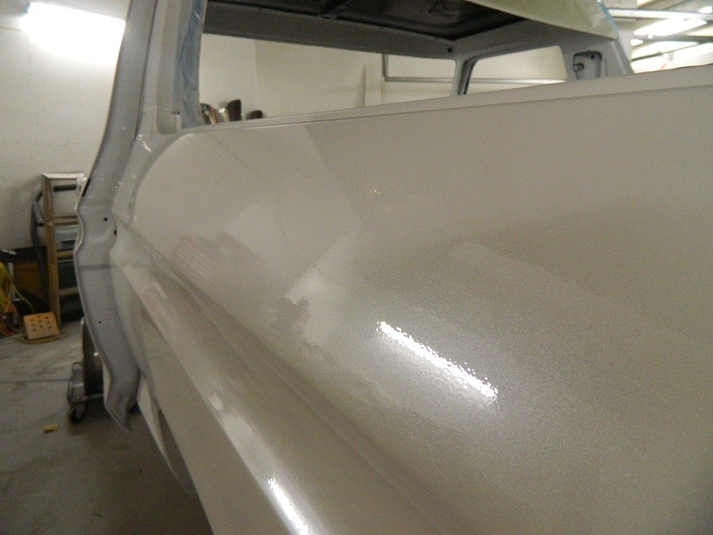 White vehicle side panel with metallic paint, showing reflections. Part of the door frame is visible