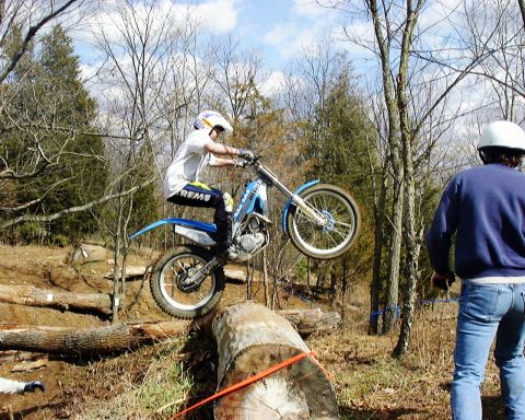 1985 Ford F-150 - A rider on a trials motorcycle navigates an obstacle course of logs in a wooded area.