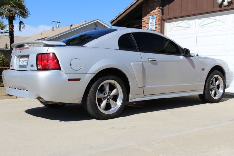 2002 Ford Mustang - A silver 2002 Ford Mustang coupe is parked in front of a garage, showing its side profile and rear.