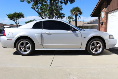 2002 Ford Mustang - A silver 2002 Ford Mustang coupe is seen from the side, parked in front of a brick garage.