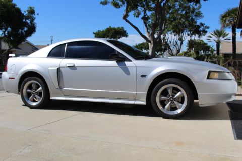 2002 Ford Mustang - A silver 2002 Ford Mustang GT coupe is shown from the side, parked on a concrete driveway with trees in the background.