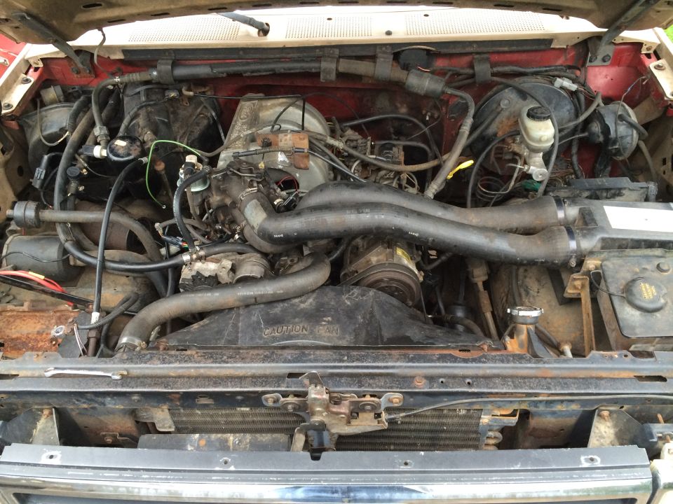 Engine bay with V8 engine, hoses, fan, and radiator visible. Caution fan warning sticker