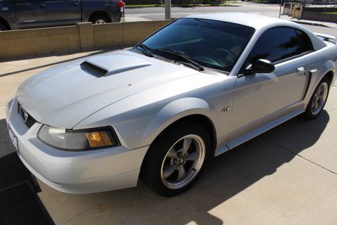 2002 Ford Mustang - A silver 2002 Ford Mustang GT is parked on a paved surface, with a dark pickup truck visible in the background.