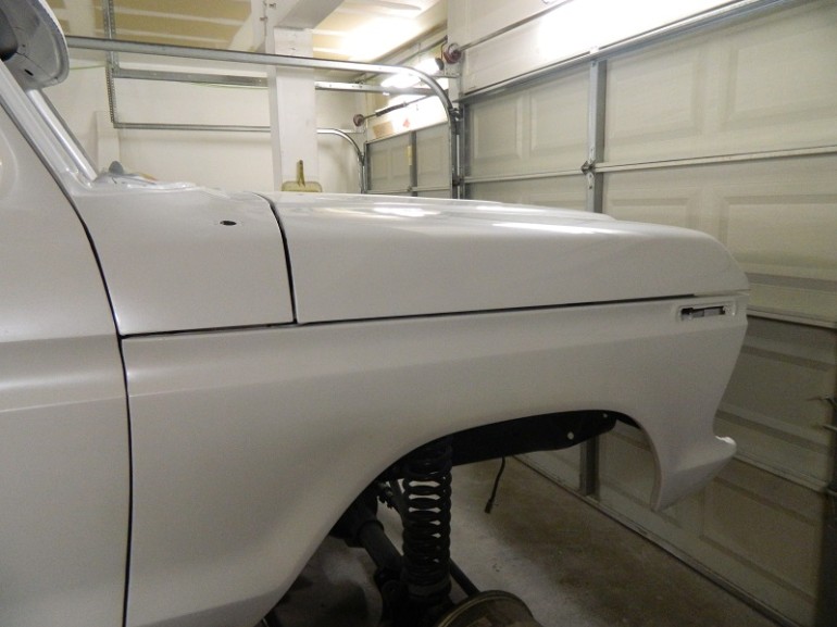 Side view of a white pickup truck fender and hood, showing suspension components. The vehicle is in a garage