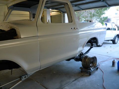 1979 Ford Bronco - A white pickup truck is being worked on in a garage, with its rear axle removed and a spare wheel nearby.