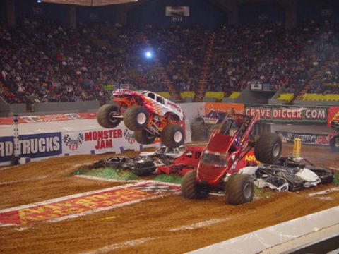 1989 Ford Bronco - Two monster trucks perform stunts over a pile of crushed cars in a crowded arena.