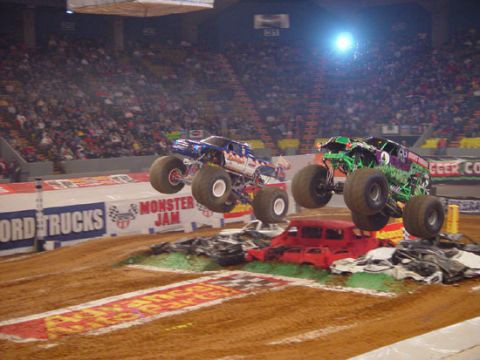 1989 Ford Bronco - Monster trucks soar over a pile of cars during a Monster Jam event, with a large crowd watching in the stadium.