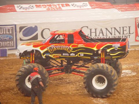 1989 Ford Bronco - A monster truck with custom paint and large tires is displayed on a dirt arena floor.