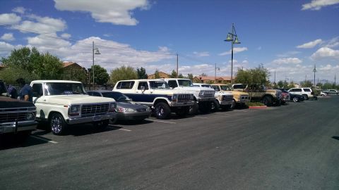 1979 Ford Bronco - A row of classic Ford trucks and SUVs are parked in a lot under a blue sky with clouds.