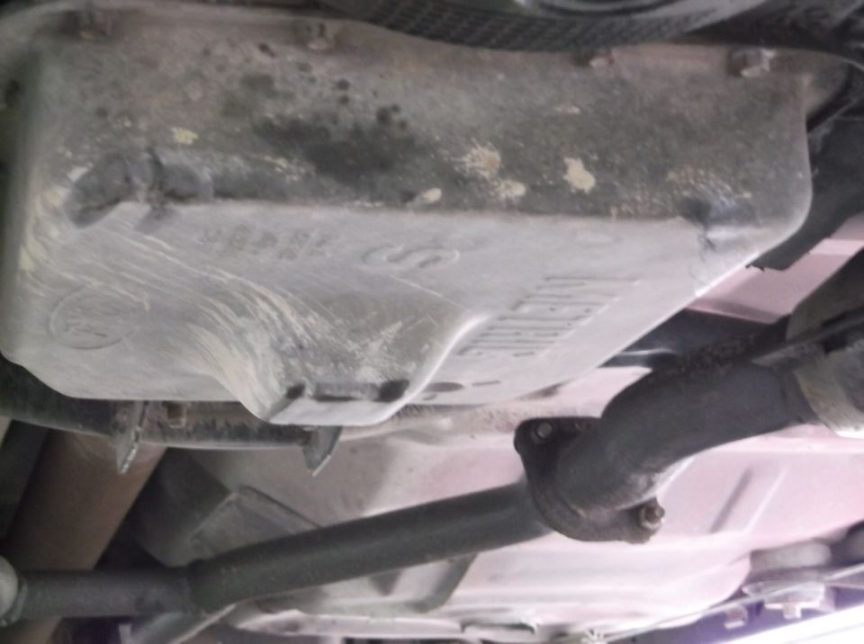 Underside view of a vehicle's oil pan and exhaust pipe. Text on the oil pan is partially visible