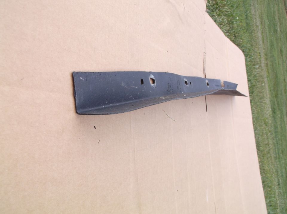 Black metal car part with holes, lying on cardboard with grass in background