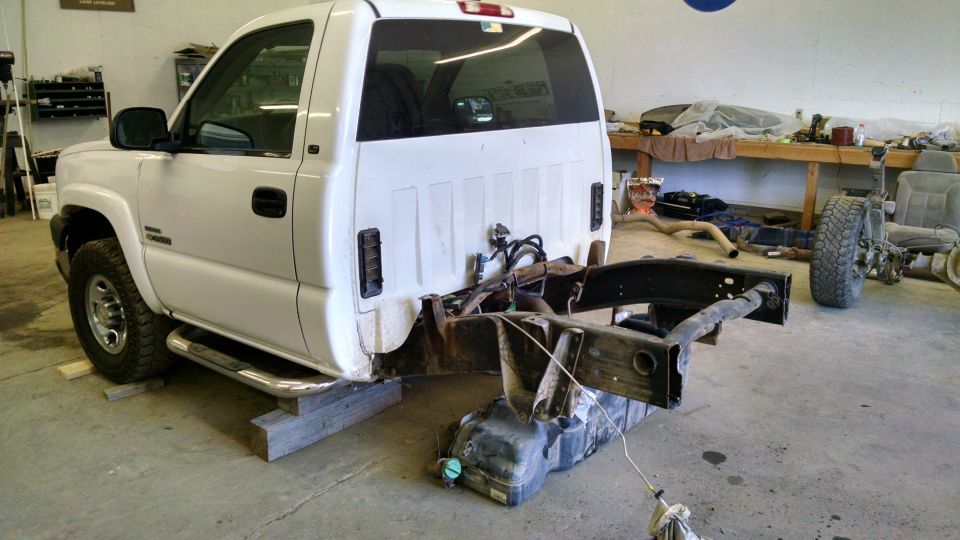 White pickup truck chassis on blocks, fuel tank below, workshop background