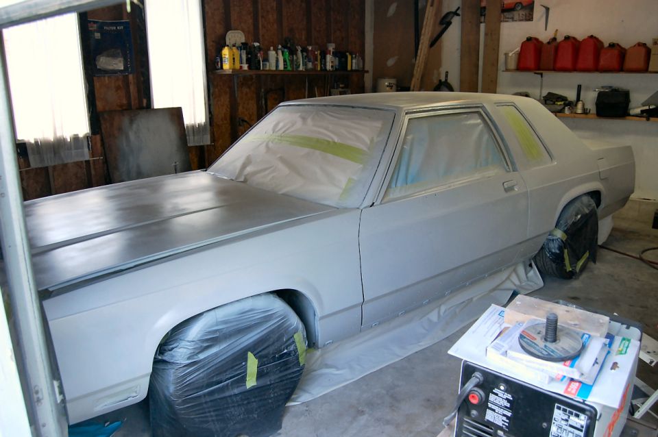 Gray sedan undergoing bodywork, covered in plastic and tape, with wheels wrapped in plastic