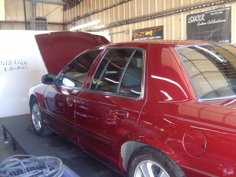 2004 Ford Crown Victoria - Red Ford full-size sedan sitting on a chassis dyno inside a metal shop with its hood raised, viewed from the driver's-side rear quarter wit…